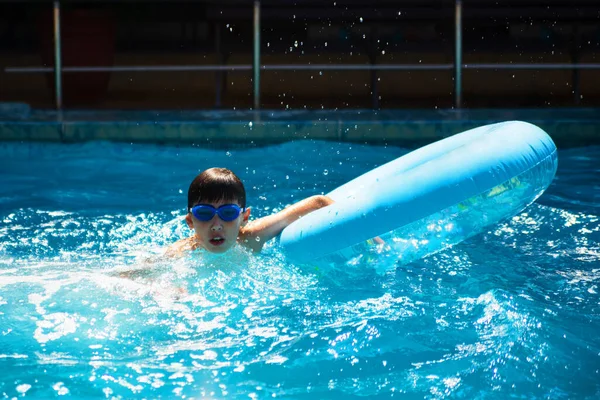 Ragazzo europeo gioca in estate in piscina nella natura. Un bambino in un cerchio gonfiabile blu spruzza gocce d'acqua. — Foto Stock