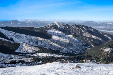 Karlı dağların geniş manzarası, ormanlı yamaçlar ve bir tatil köyü.