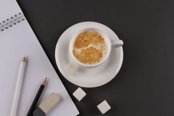 A white coffee Cup and a drawing pad with pencils on a gray background