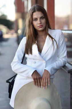 Modern bir şehrin arka planında beyaz ceketli ve yaz şapkalı bir kızın portresi.