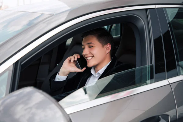 ¡Hablando mientras conduces! Joven sonriente sentado al volante de su coche conduciendo por la ciudad y comunicándose por teléfono inteligente . — Foto de Stock