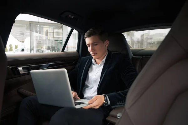 Ήρθε η ώρα να εργαστούν! Όμορφος όμορφος νεαρός άνδρας απασχολημένος σοβαρά να κάνει τη δουλειά του για το φορητό υπολογιστή στο πίσω κάθισμα στο αυτοκίνητο. — Φωτογραφία Αρχείου