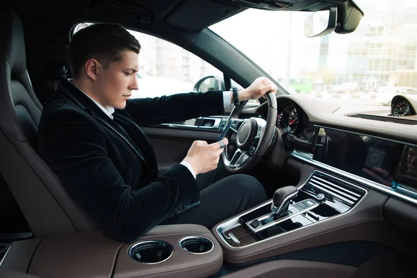 Mensajes de texto mientras conduce! Retrato de vista lateral del joven hombre de negocios guapo confiado que fija su teléfono inteligente, mientras que conduce el coche . — Foto de Stock