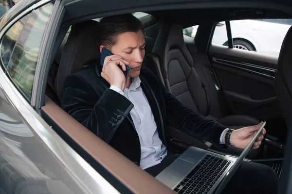¡Mantente siempre disponible! Joven hombre educado bien parecido sentado en el coche, hablando el teléfono y trabajando en su computadora inalámbrica . — Foto de Stock
