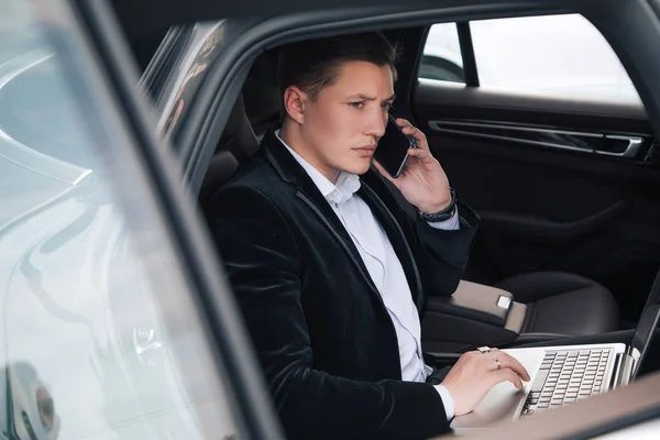 ¡Habla de negocios! Siempre en línea joven serio guapo recién llegado trabajando en su ordenador portátil en el coche y hablando de su teléfono inteligente . — Foto de Stock
