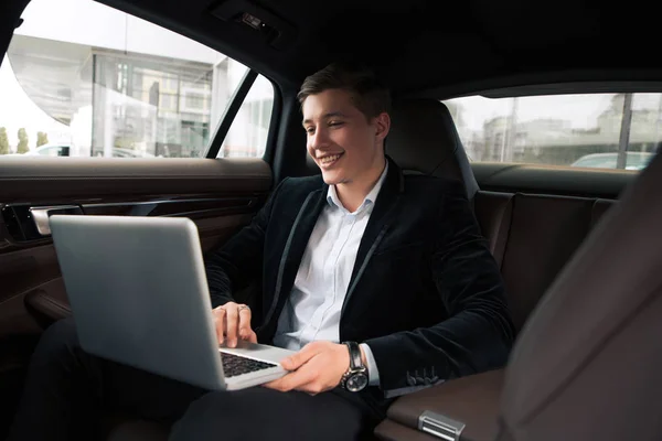 Τη δουλειά με ευχαρίστηση! Όμορφος χαμογελαστός νεαρός άνδρας που κάνει τις επιχειρήσεις στο πίσω κάθισμα του αυτοκινήτου του, ενώ χρησιμοποιείτε τον ασύρματο υπολογιστή. — Φωτογραφία Αρχείου