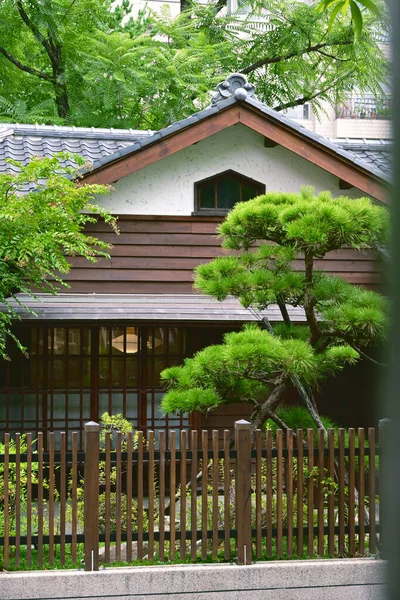 Geleneksel Japon mimarisinde koyu ahşap alt duvarlar, beyaz alçı üst kısım ve koyu kiremit çatı, ön bahçesinde zarif bir çam ağacı dikey ahşap çit arkasında yer almaktadır..