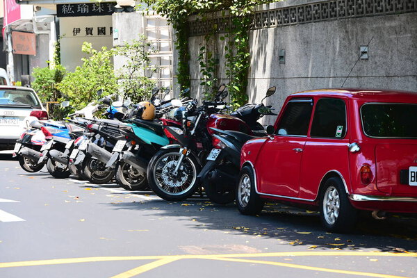 Taiwan - Sep 14, 2025: Red classic car parked roadside with five motorcycles in various colors including blue, black and silver, on asphalt road with yellow markings and building background.