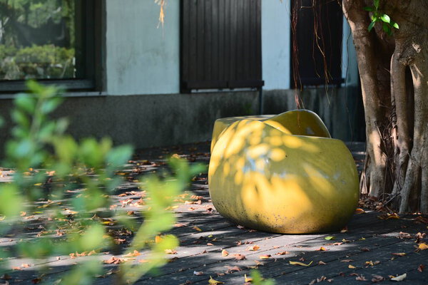 Большая жёлтая изогнутая скульптурная инсталляция сидений в общественном парке с пышными тенями деревьев, естественной листвы и традиционным архитектурным фоном.