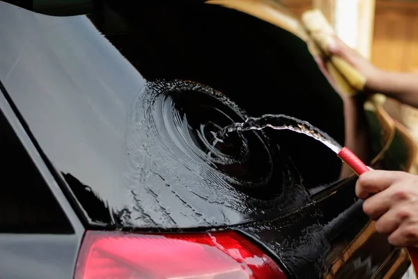 O homem lava um carro preto com uma mangueira. Lavagem de auto-carro. — Fotografia de Stock