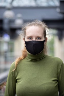 Güzel mimarisi olan bir binanın içinde duran siyah maskeli güzel bir kadının portresi. Koyu yeşil kazaklı ve haki pantolonlu bir kadın..