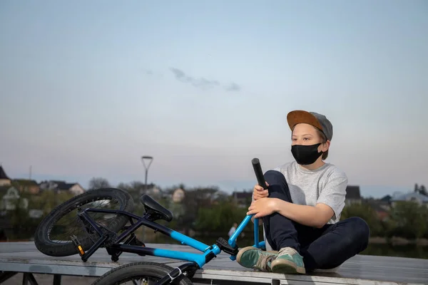 Portrait of a teenager in the sunset in a black mask on a blue bicycle. Quarantine. It is forbidden to ride a bicycle. Covid-19.