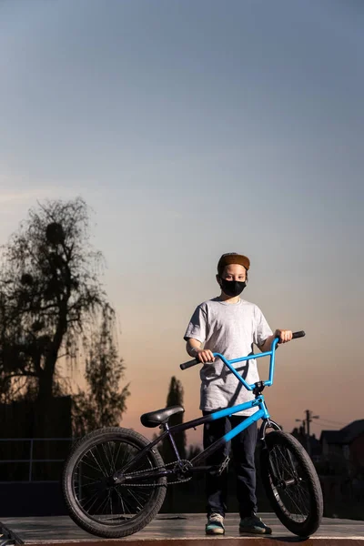 Portrait of a teenager in the sunset in a black mask on a blue bicycle. Quarantine. It is forbidden to ride a bicycle. Covid-19.