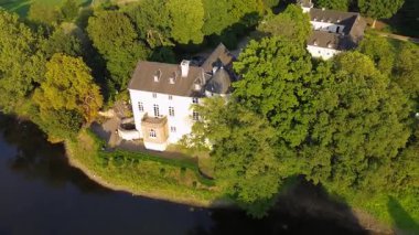 Sakin bir nehrin yakınında bulunan yemyeşil ormanlarla çevrili tarihi beyaz bir kale. Hava manzarası, temiz bir gökyüzünün altında ağaçlar, tarlalar ve uzak köylerle huzurlu bir kırsal bölgeyi vurguluyor..