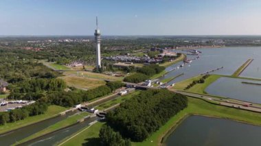 Yüksek bir iletişim kulesi nehir kenti üzerinde yükselmekte, kanallar, köprüler, botlar ve yeşil manzaralarla modern altyapıyı doğal çevreyle harmanlamaktadır..