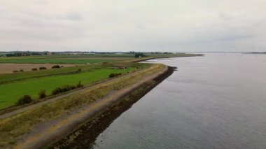 Yeşil tarlaları, dolambaçlı yolları ve kıyı boyunca uzanan sakin bir kıyı manzarası bulutlu bir gökyüzünün altında karanın ve denizin barışçıl uyumunu gösteren bir deniz manzarası..