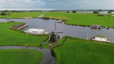 Uçsuz bucaksız yeşil çayırlar ve Hollanda bayraklarıyla kesişen, bulutlu bir gökyüzünün altında rüzgarda hafifçe sallanan su yollarıyla çevrili, nehirde yüzen evlerin sakin bir görüntüsü. Klasik bir Hollanda yel değirmeni