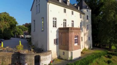 Sakin bir nehrin yakınında bulunan yemyeşil ormanlarla çevrili tarihi beyaz bir kale. Hava manzarası, temiz bir gökyüzünün altında ağaçlar, tarlalar ve uzak köylerle huzurlu bir kırsal bölgeyi vurguluyor..