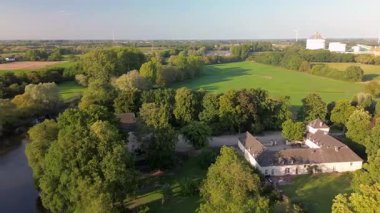 Bereketli yeşil tarlalar, bir nehir ve yüksek bacaları ve rüzgâr türbinleri olan endüstriyel bir bitki. U şeklinde avlusu olan tarihi bir malikane..