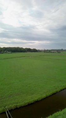 Geniş yeşil alanlar, dolambaçlı bir kanal ve bulutlu bir gökyüzünün altında yağmurdan önce huzurlu kırsal atmosferi yakalayan uzak bir köy..