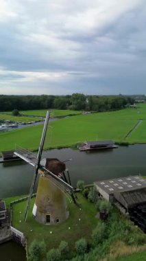 Çiçekli yeşil tarlalar, geleneksel evler ve bulutlu bir gökyüzünün altındaki uzak su yollarıyla çevrili bir kanalın yanındaki Hollandalı yel değirmeni manzarası..