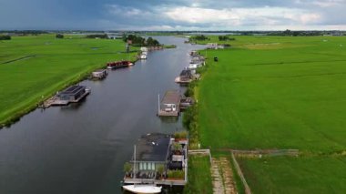 Uçsuz bucaksız yeşil çayırlar ve Hollanda bayraklarıyla kesişen, bulutlu bir gökyüzünün altında rüzgarda hafifçe sallanan su yollarıyla çevrili, nehirde yüzen evlerin sakin bir görüntüsü. Klasik bir Hollanda yel değirmeni