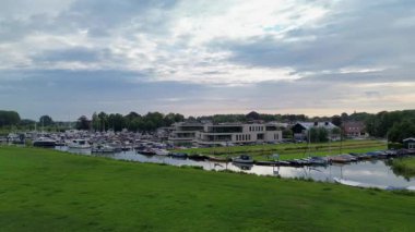 Yeşil tarlalar, modern binalar ve bulutlu bir gökyüzünün altındaki köyle çevrili nehir boyunca tekneleri ve yatları olan huzurlu bir marina..