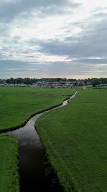 Sarmal bir dere yemyeşil çayırlar boyunca akar, yukarıdaki bulutlu gökyüzünü yansıtır. Uzakta, binalar ve bir marina kasabanın kenarında, huzurlu bir kırsal alan yaratıyor..