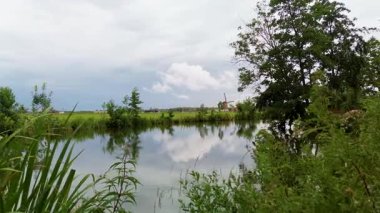 Bereketli yeşilliklerle çevrili, gökyüzünü yansıtan sakin bir nehri olan huzurlu kırsal manzara. Geleneksel bir yel değirmeni uzakta duruyor ve kırsal alana cazibe katıyor..