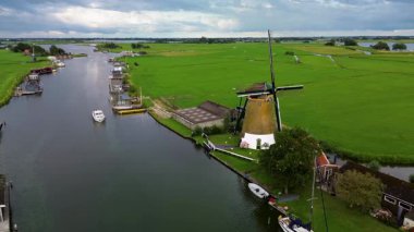 Huzurlu bir nehir manzarası. Yeşil tarlalardan, geleneksel evlerden ve kırsal kesimin cazibesini yakalayan tarihi yel değirmeninden sakin bir şekilde geçen tekneler..
