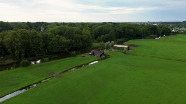 Dolambaçlı bir dere geniş yeşil çayırlar boyunca akar, orman kenarındaki küçük kırsal ahırlar ve sakin, bulutlu bir gökyüzü altında uzak bir kasaba.