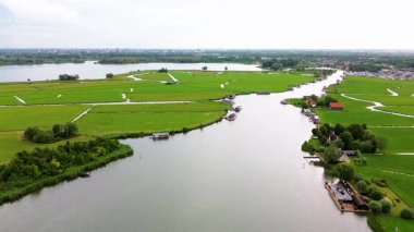 Huzurlu bir nehir manzarası. Yeşil tarlalardan, geleneksel evlerden ve kırsal kesimin cazibesini yakalayan tarihi yel değirmeninden sakin bir şekilde geçen tekneler..