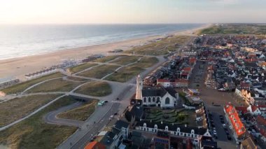 Gün batımında deniz kenarı manzarası, kırmızı çatılı evler, tarihi bir kilise, kumlu kumullar, sahil kulübeleri ve altın gece ışığı altında parlayan sakin deniz..