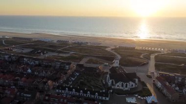 Gün batımında deniz kenarı manzarası, kırmızı çatılı evler, tarihi bir kilise, kumlu kumullar, sahil kulübeleri ve altın gece ışığı altında parlayan sakin deniz..