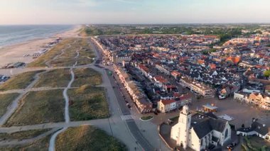 Gün batımında deniz kenarı manzarası, kırmızı çatılı evler, tarihi bir kilise, kumlu kumullar, sahil kulübeleri ve altın gece ışığı altında parlayan sakin deniz..