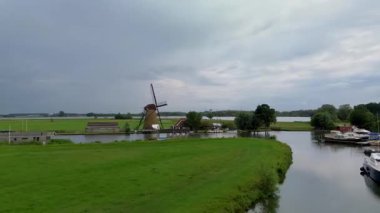Huzurlu bir nehir manzarası. Yeşil tarlalardan, geleneksel evlerden ve kırsal kesimin cazibesini yakalayan tarihi yel değirmeninden sakin bir şekilde geçen tekneler..