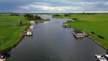 Huzurlu bir nehir manzarası. Yeşil tarlalardan, geleneksel evlerden ve kırsal kesimin cazibesini yakalayan tarihi yel değirmeninden sakin bir şekilde geçen tekneler..