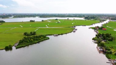 Huzurlu bir nehir manzarası. Yeşil tarlalardan, geleneksel evlerden ve kırsal kesimin cazibesini yakalayan tarihi yel değirmeninden sakin bir şekilde geçen tekneler..