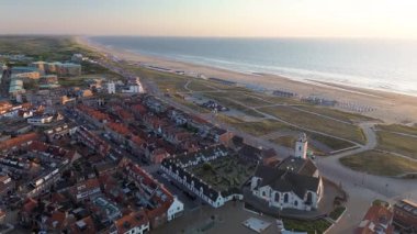 Gün batımında deniz kenarı manzarası, kırmızı çatılı evler, tarihi bir kilise, kumlu kumullar, sahil kulübeleri ve altın gece ışığı altında parlayan sakin deniz..