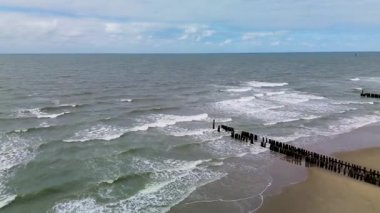 Sakin bir sahil sahnesi, yumuşak dalgalar, denize uzanan ahşap dalgalar, kumsalda yürüyen ve balık tutan birkaç insan, ve kıyıya yakın park etmiş yalnız bir araba..