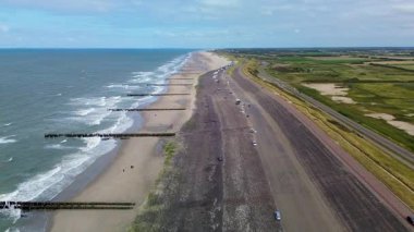 Okyanus dalgaları, dalgalar, park etmiş arabalar ve ufuk boyunca uzanan geniş bir kıyı yolu olan kumlu bir sahil manzarası yeşil tarlalar ve açık bir manzara..