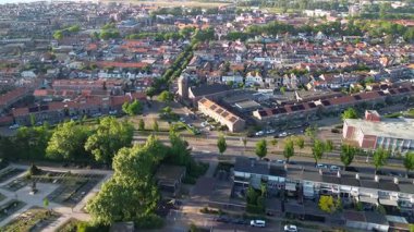 Modern evler, yeşil bahçeler ve ağaçlarla kaplı caddelerle dolu bir banliyö manzarası. Arka planda kırmızı çatıları olan yoğun bir kasaba ve bir mezarlık görülebilir.