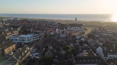 Gün batımında deniz kenarı manzarası, kırmızı çatılı evler, tarihi bir kilise, kumlu kumullar, sahil kulübeleri ve altın gece ışığı altında parlayan sakin deniz..