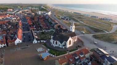 Gün batımında deniz kenarı manzarası, kırmızı çatılı evler, tarihi bir kilise, kumlu kumullar, sahil kulübeleri ve altın gece ışığı altında parlayan sakin deniz..