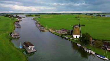 Huzurlu bir nehir manzarası. Yeşil tarlalardan, geleneksel evlerden ve kırsal kesimin cazibesini yakalayan tarihi yel değirmeninden sakin bir şekilde geçen tekneler..