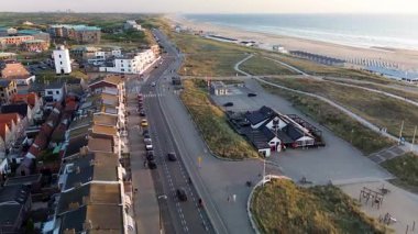 Gün batımında deniz kenarı manzarası, kırmızı çatılı evler, tarihi bir kilise, kumlu kumullar, sahil kulübeleri ve altın gece ışığı altında parlayan sakin deniz..