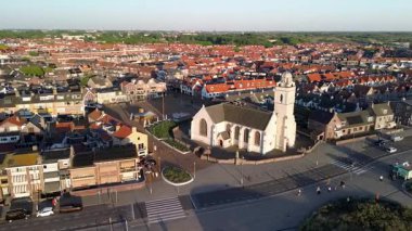 Merkezi beyaz bir kilise ve saat kulesi olan, kırmızı çatılı binalar, dar sokaklar ve kaldırımlı bir meydanla çevrili bir kasabanın hava manzarası. Tarihin ve modern şehir planının bir karışımı..