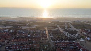 Gün batımında kıyı kasabasının havadan görünüşü, beyaz bir kilise, kırmızı çatılı evler, çimenli kumullar ve plaj kabinleri. Altın gökyüzü ve sakin okyanus huzurlu, resim gibi bir atmosfer yaratır..