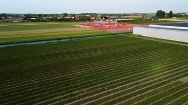Canlı çiçek sıraları ve yürüyen ziyaretçilerle bir kanala bölünmüş tarım arazilerinin havadan görünüşü. Arka plandaki seralar ve kırsal binalar tarım ve turizm entegrasyonunu ön plana çıkarıyor.