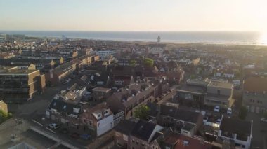 Gün batımında kıyı kasabasının havadan görünüşü, beyaz bir kilise, kırmızı çatılı evler, çimenli kumullar ve plaj kabinleri. Altın gökyüzü ve sakin okyanus huzurlu, resim gibi bir atmosfer yaratır..